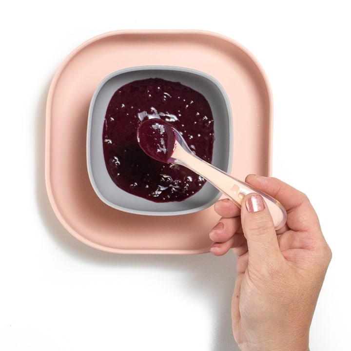 Pink baby plate with gray bowl full of blueberry puree with hand holding pink baby spoon full of puree.