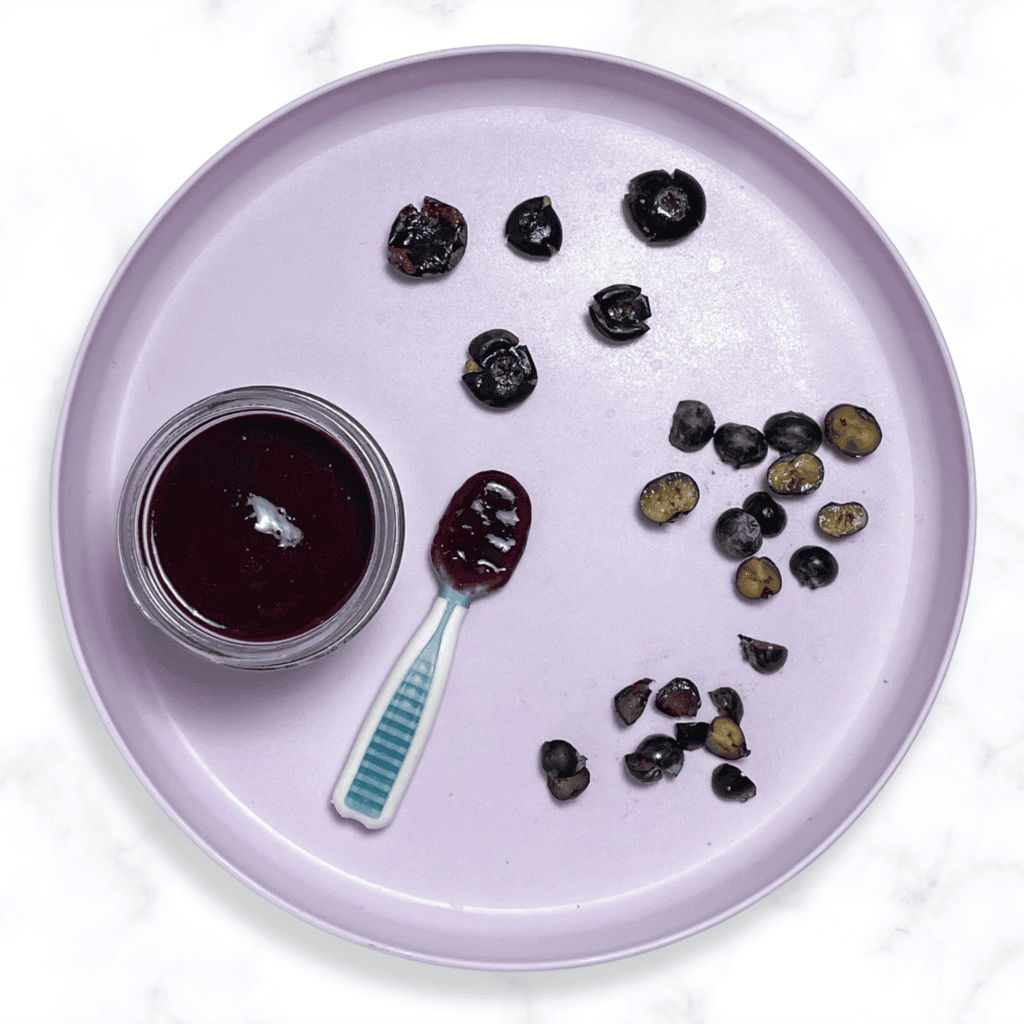 Purple baby plate with self-feeding spoons showing different ways to serve blueberries to baby. The baby plate is sitting on my marble kitchen counter. 
