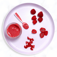 Purple baby plate sitting on a marble counter. On the plate is a raspberry puree and pink baby spoon as well as options on how to cut and serve raspberries as a finger food.