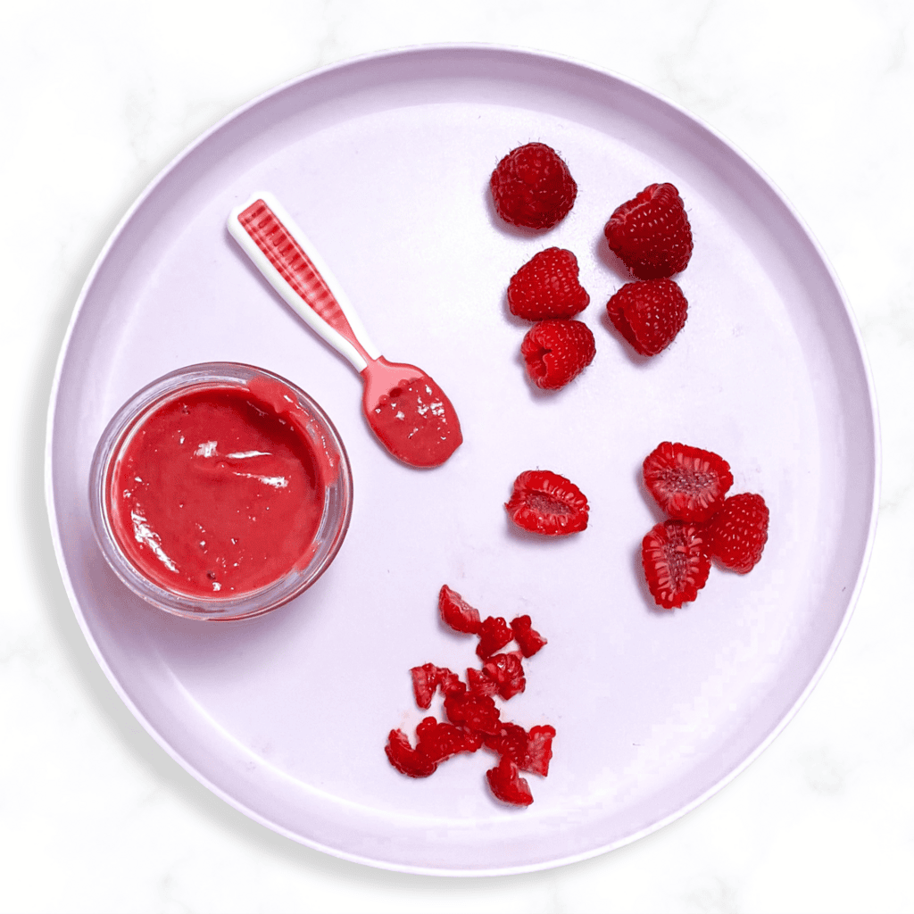 Purple baby plate showing different ways to serve raspberries to your baby - as a puree with a pink spoon or in different finger food sizes. 