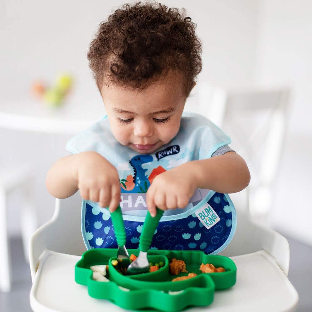 Boy sitting in white hair chair eating out of a green plate with green utensils