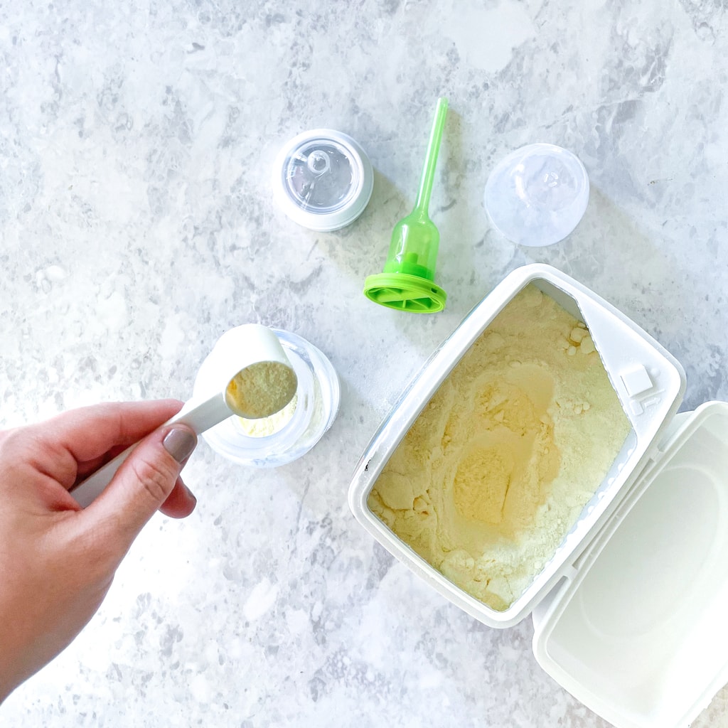 A bottle on a marble countertop with a jar of baby formula with a hand pouring the baby formula in the bottle.