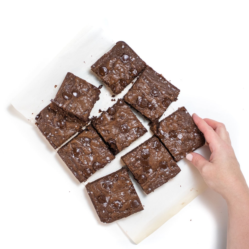 A hand holding a brownie and a grid of nine brownies on a white parchment paper against a white background.
