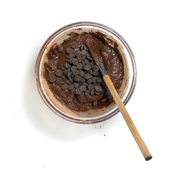 A glass mixing bowl with the wet and dry ingredients combined with chocolate chips on top and a spatula ready to mix.