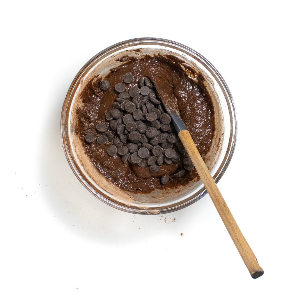 A glass mixing bowl with the wet and dry ingredients combined with chocolate chips on top and a spatula ready to mix.