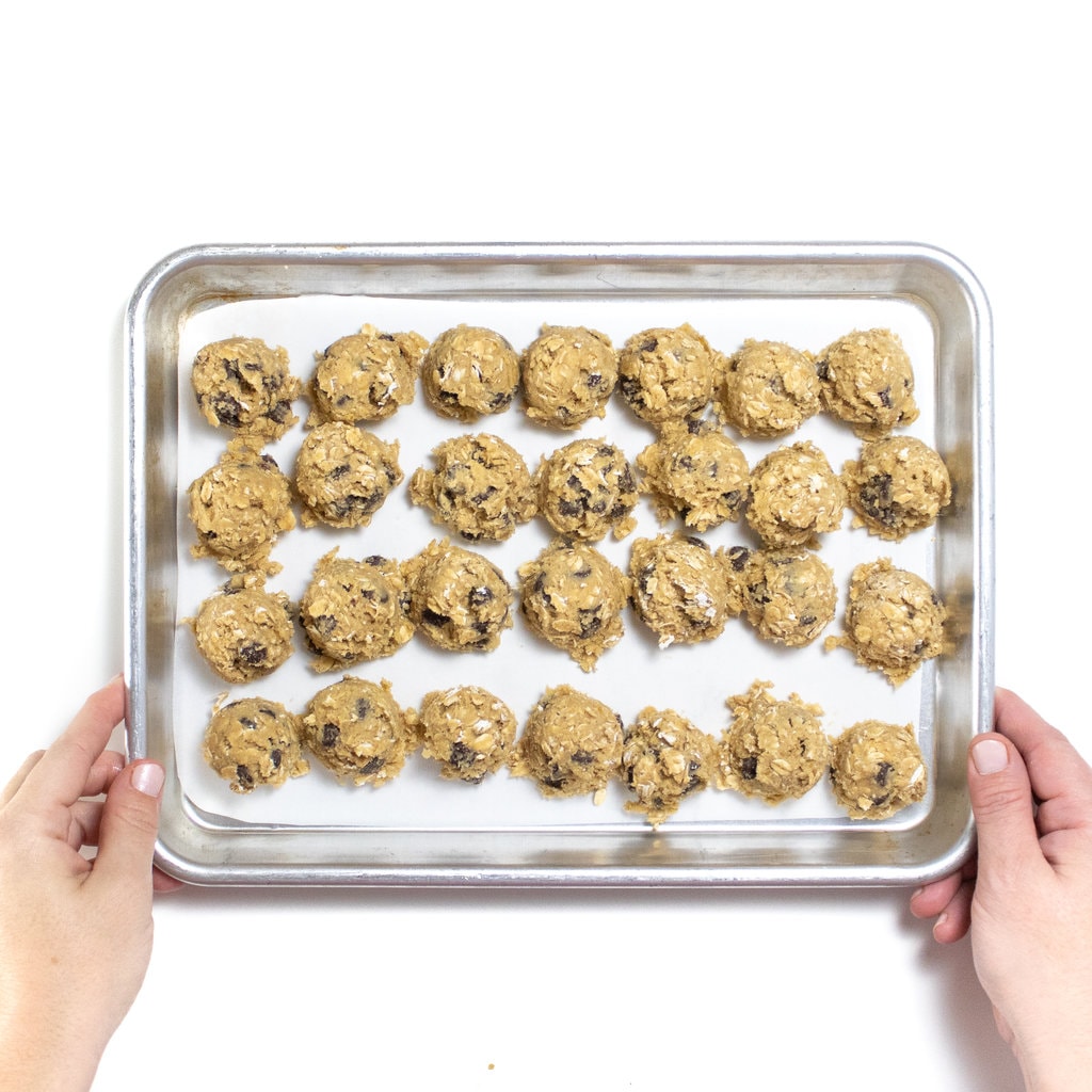 Two hands holding a silver baking sheet full of scoops of cookie dough ready for the freezer.
