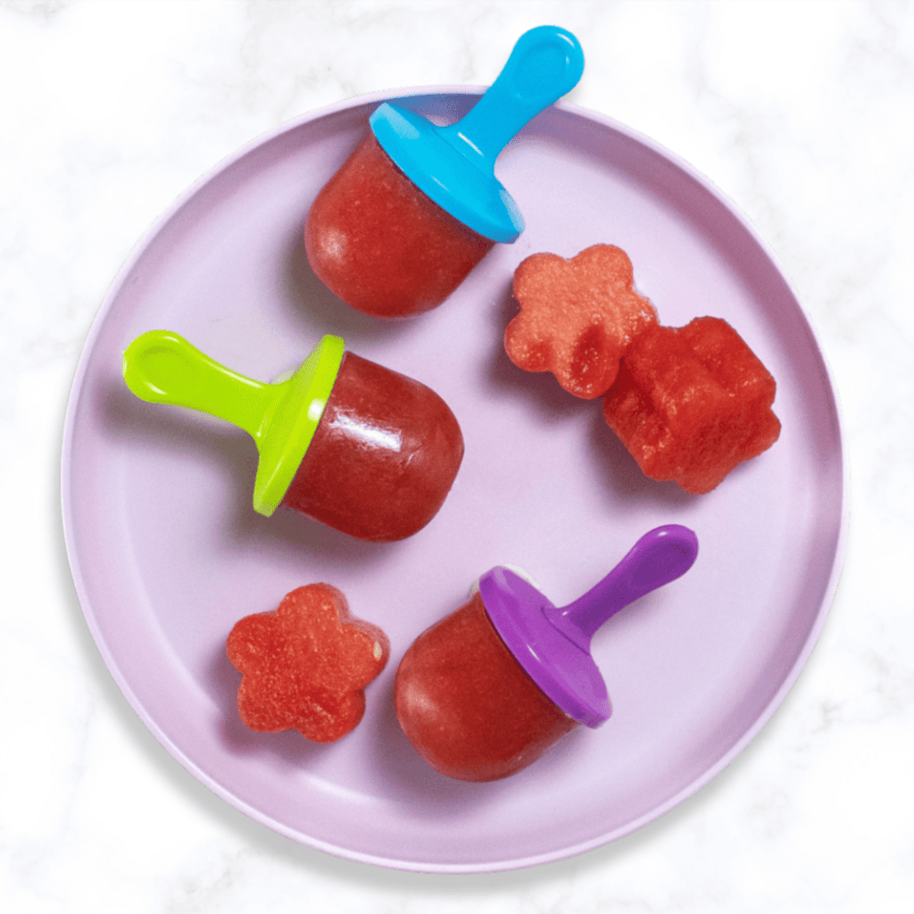 purple kids plate with 3 small watermelon popsicles and a few fun cut out shapes of watermelon. The plate is sitting on my white marble kitchen counter. 