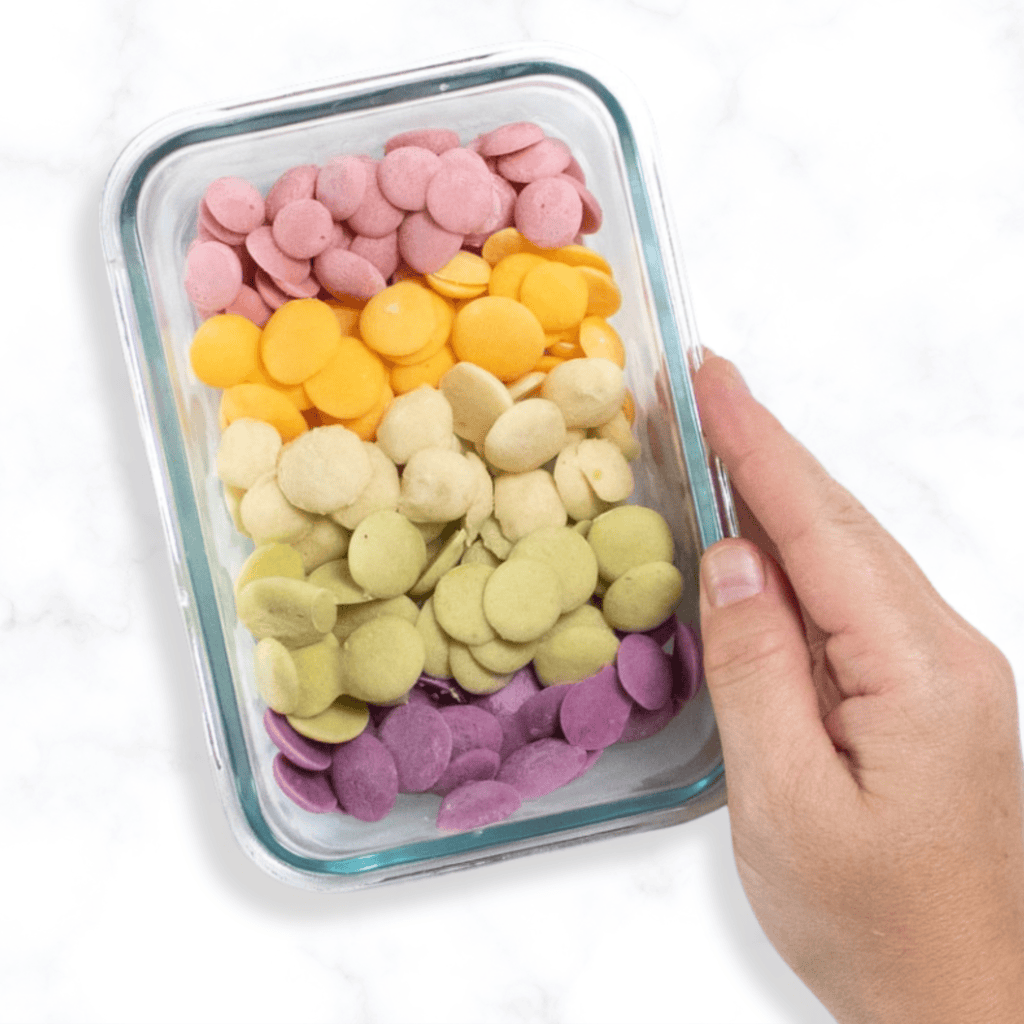 Hand holding a small glass container on a marble kitchen countertop, with several different flavors of yogurt melts for baby. 