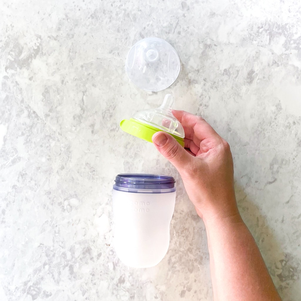 Comotomo bottles taken apart on a marble countertop with a hand holding a lid.