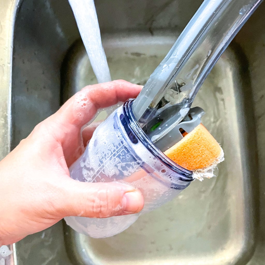 A hand holding a comotomo baby bottle inside a sink washing it was a dish brush.