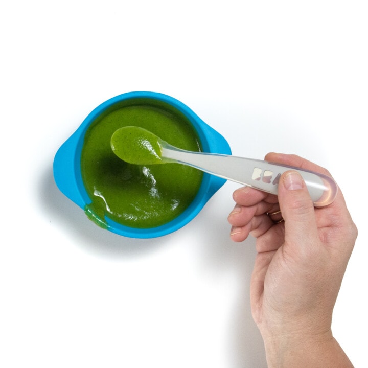 A blue baby bowl filled with a smooth zucchini purée with a hand holding a spoon.
