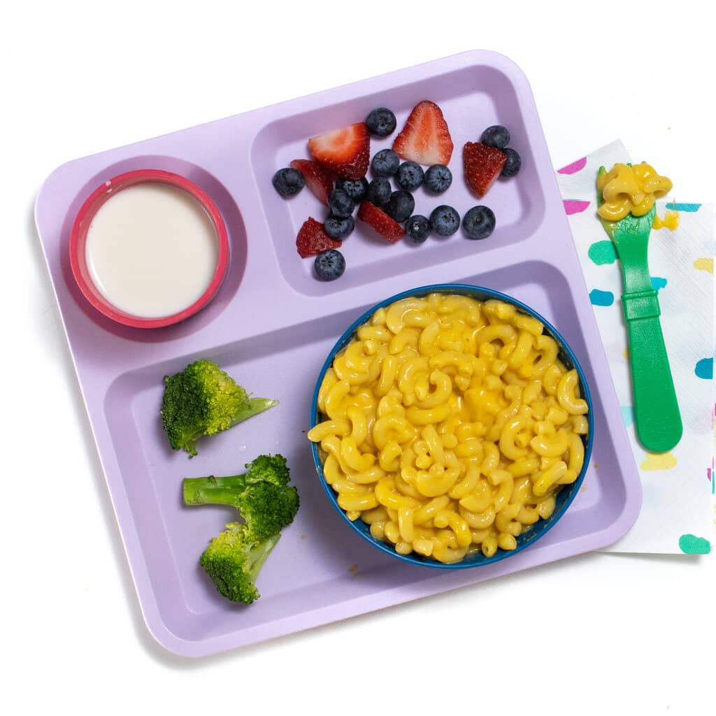 A purple kids school tray for lunch filled with a blue bowl full of butternut squash mac & cheese, broccoli, berries in milk.