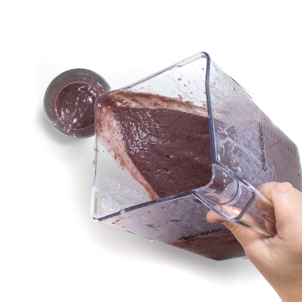 A hand holding a blender pouring a pregnancy smoothie into a clear glass against a white background.