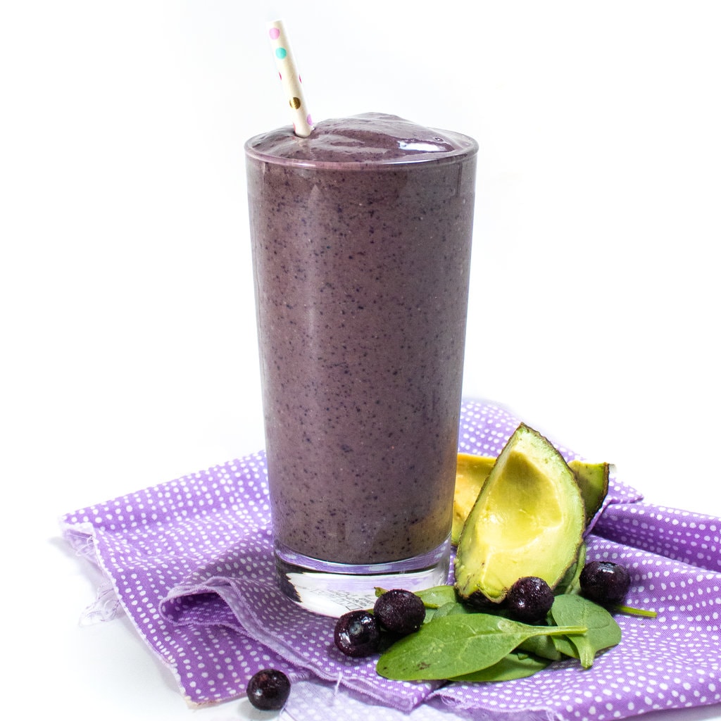 A side view of a pregnancy smoothie that is purple with a purple napkin and ingredients scattered at the bottom.