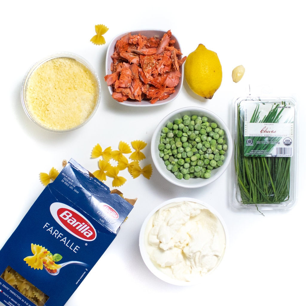 Spread of ingredients needed for this recipe against a white background - pasta, peas, salmon, ricotta, chives, lemon, garlic, and parmesan.