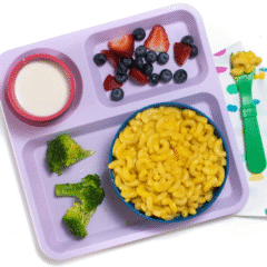 My hand holding a green kids spoon over a bowl of creamy butternut squash mac and cheese. The mac and cheese is in a blue kids bowl sitting on a school purple plate with a side of broccoli and fruit, all sitting on my white marble kitchen counter.