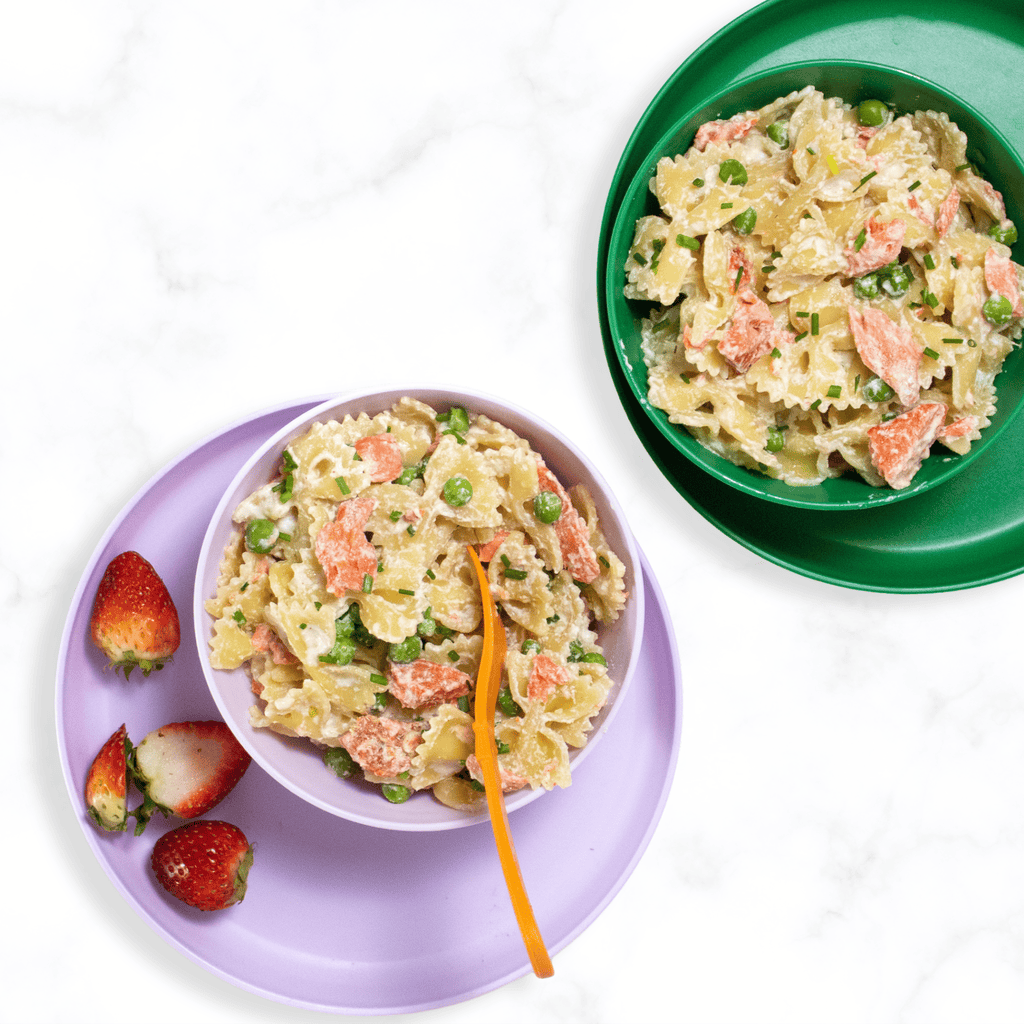 Two colorful kids bowls on plates sitting on a marble white kitchen counter filled with pasta, salmon and peas. 