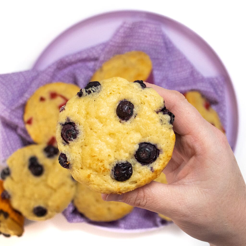 A hand holding a blueberry yogurt muffin over a plate on my white countertop in my kitchen.