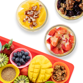 Three yogurt parfaits in a jar with a tray next to it full of different kinds of toppings all kid friendly.