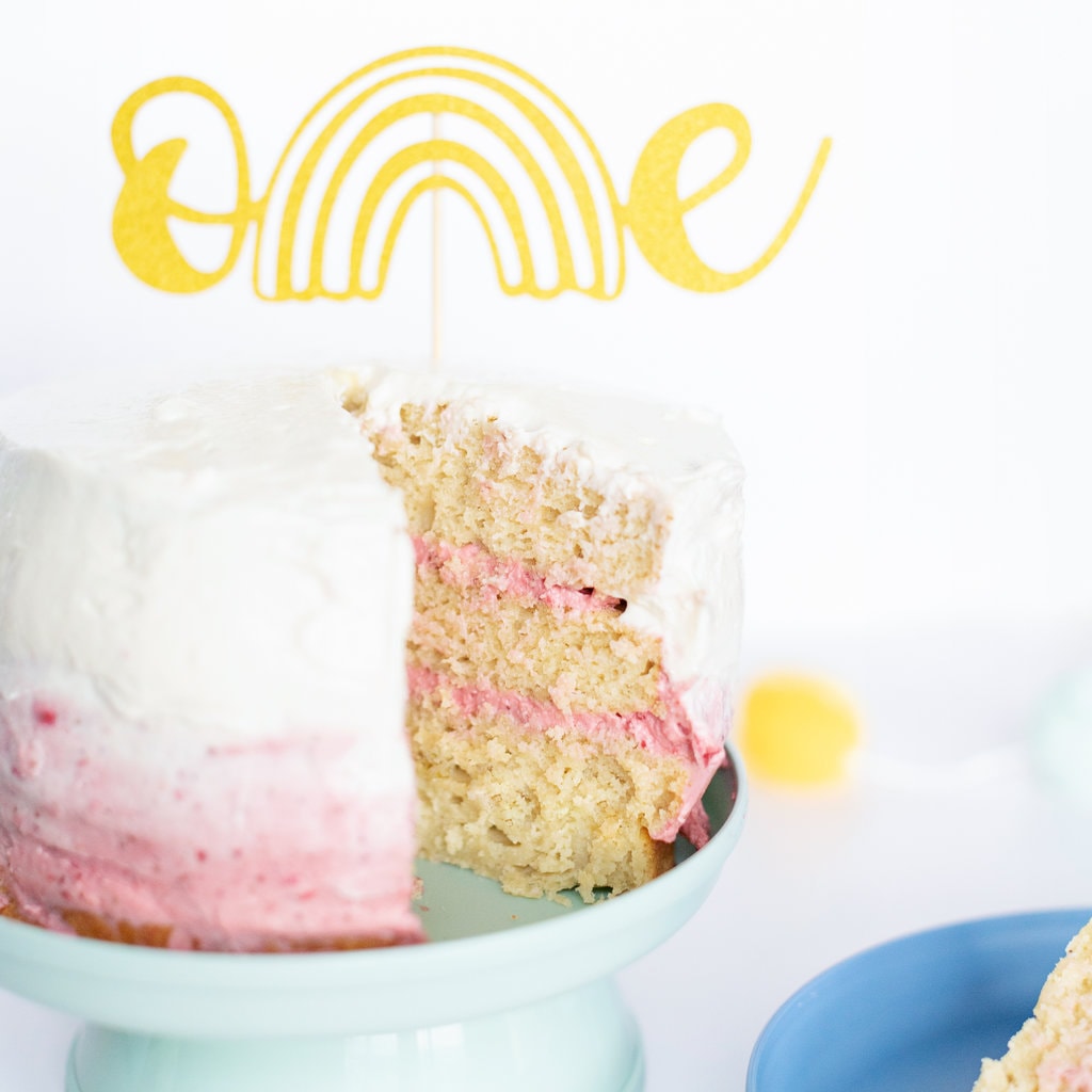 Smash cake with pink and white frosting on a teal cake stand with a slice of cake next to it on a blue plate.