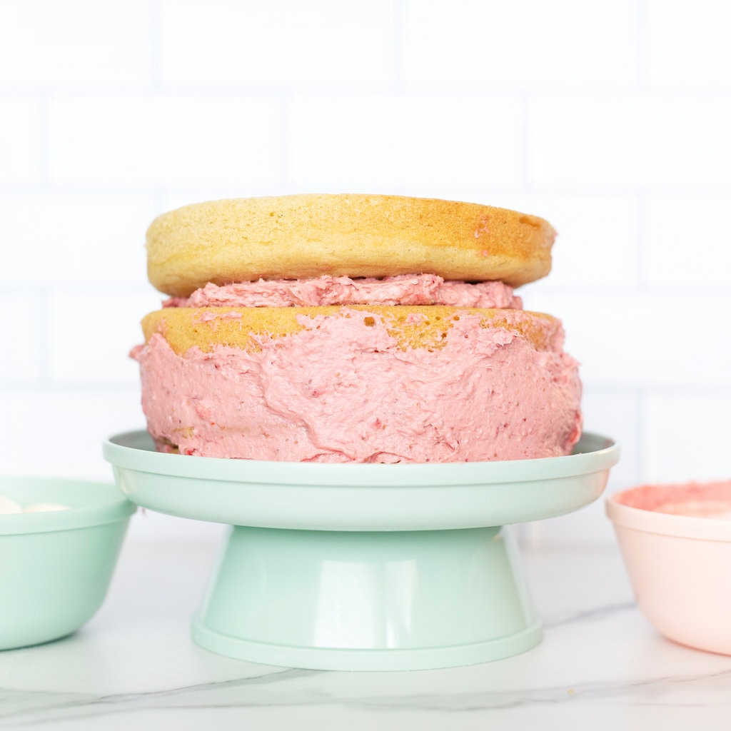 A smash cake for baby getting assembled on a teal cake stand with pink healthy cream cheese icing.