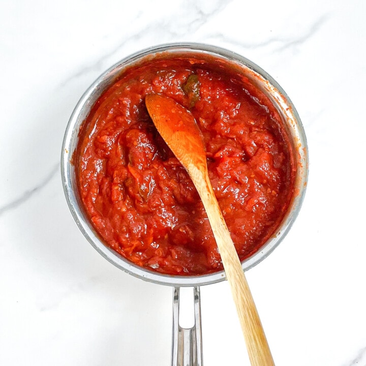 Silver saucepan on a marble surface with cooked down tomatoes and veggies for a pizza sauce.