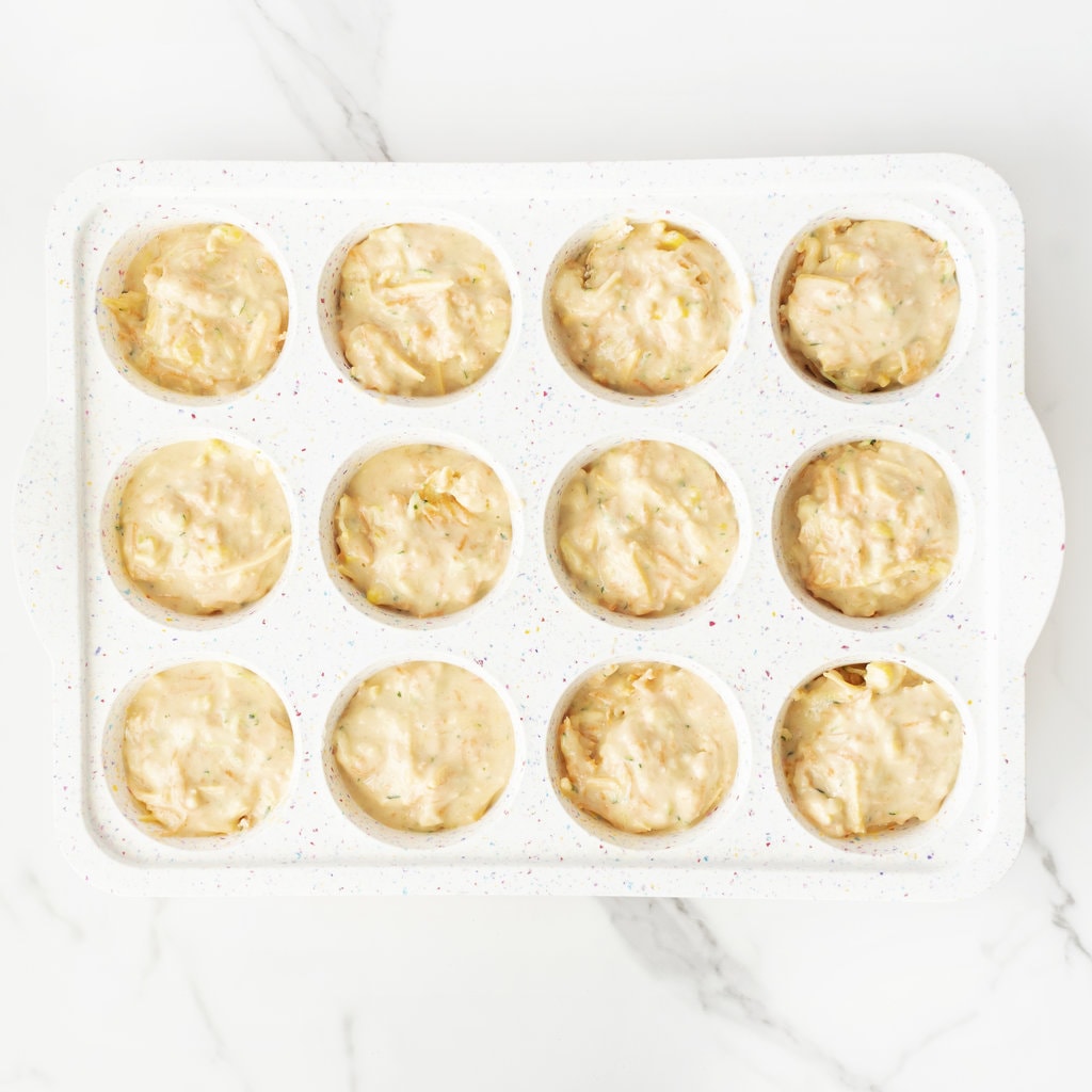 A muffin tray full of veggie muffins ready to be baked on a marble countertop.