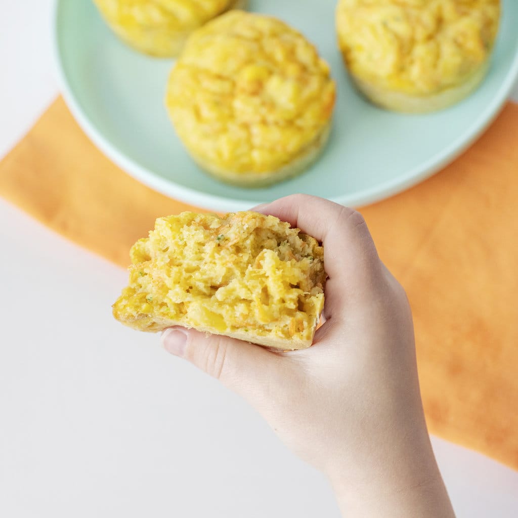 Hey kids hand holding a veggie muffin torn in half against a background of a blue kids plate and orange napkin.