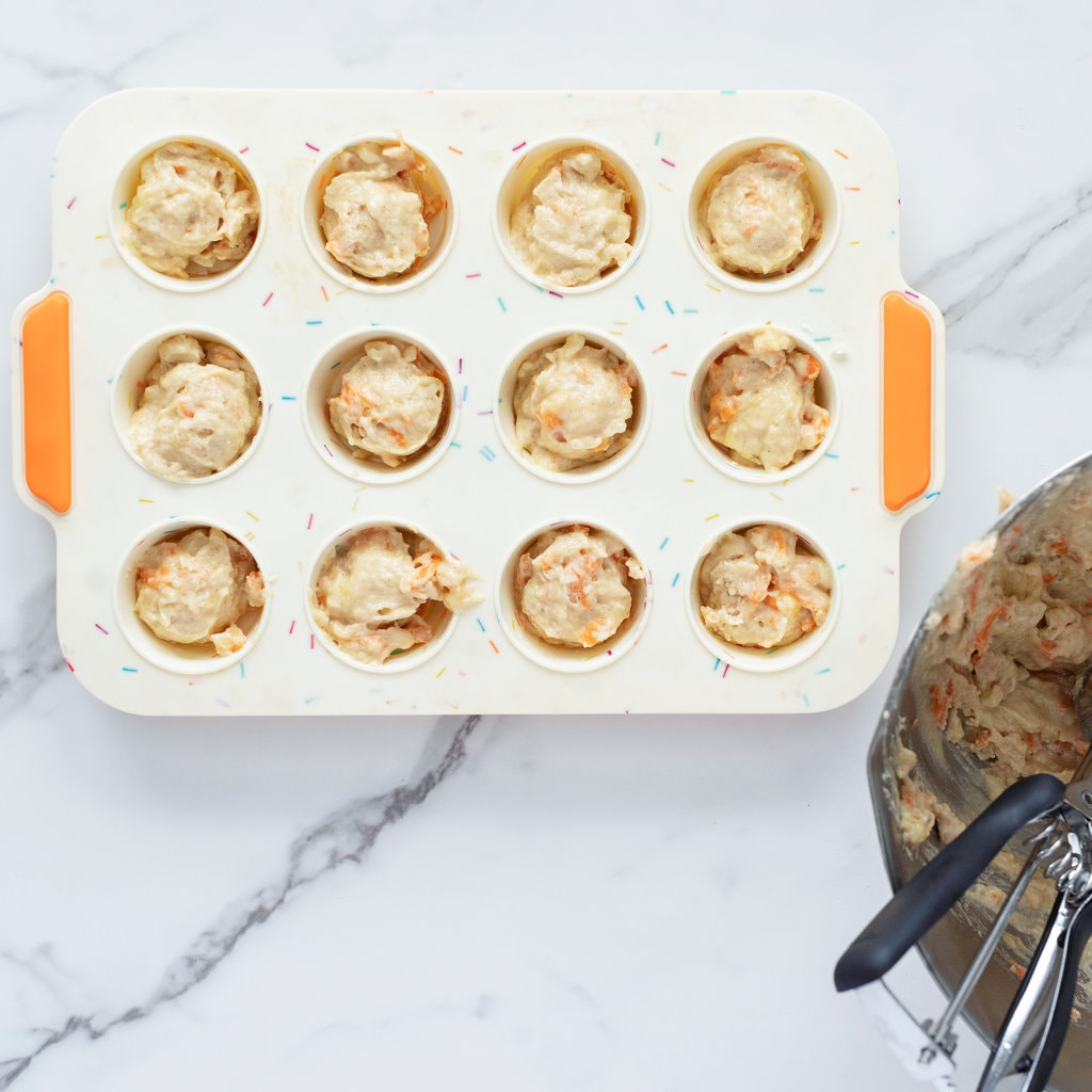A colorful muffin pan been filled with the batter of muffins for baby and toddler on a marble countertop.