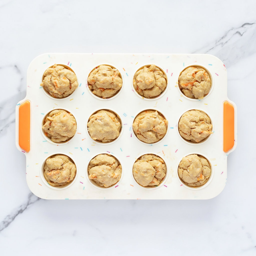 A colorful muffin tray with ABC muffins made with apple, banana and carrots, sitting on a marble countertop.