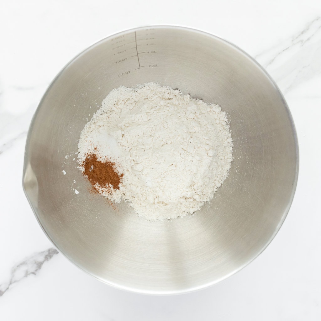 Silver mixing bowl with flour, baking powder, salt and cinnamon on a marble counter.