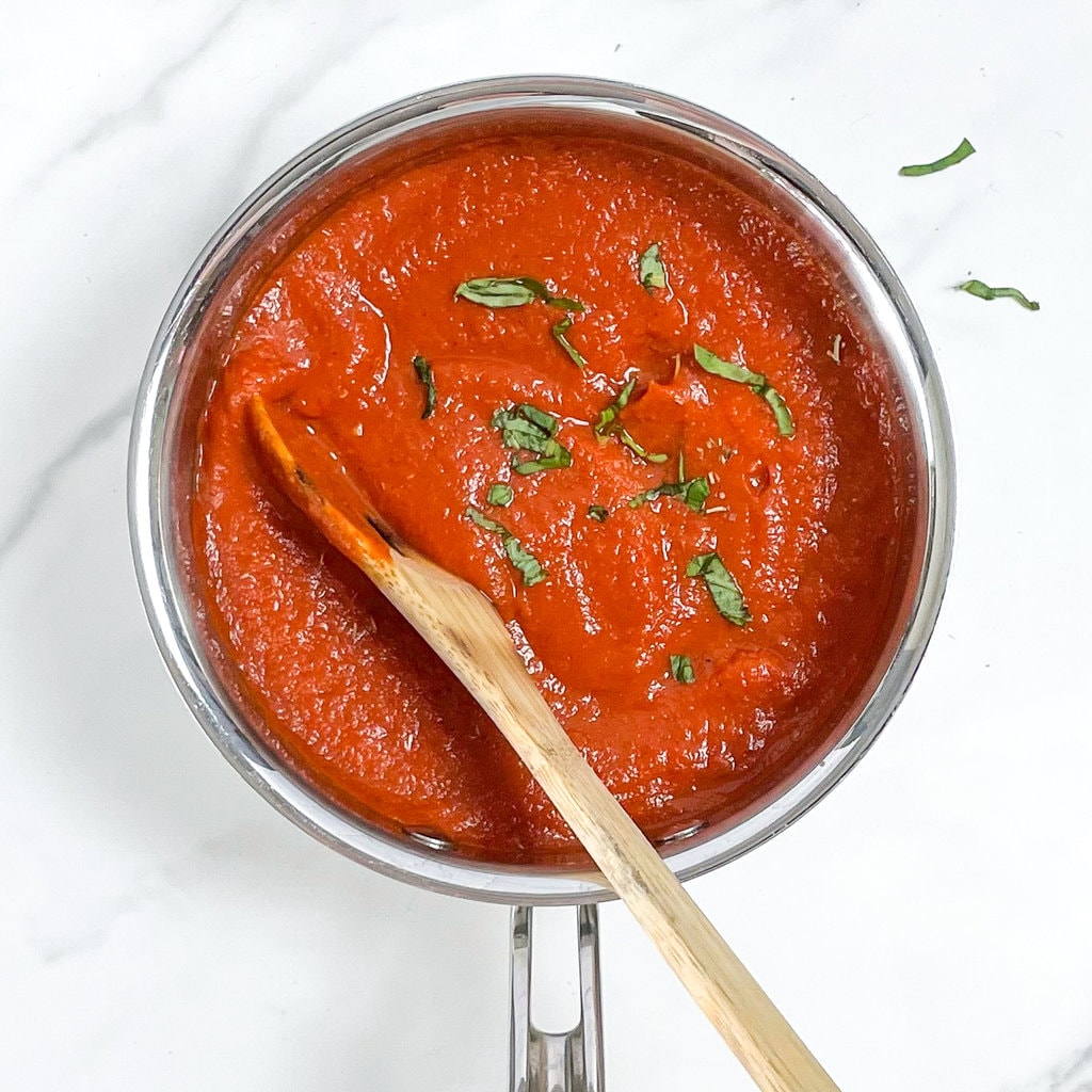 Silver saucepan on a marble counter with a red pizza sauce, basil and a wood spoon resting inside.