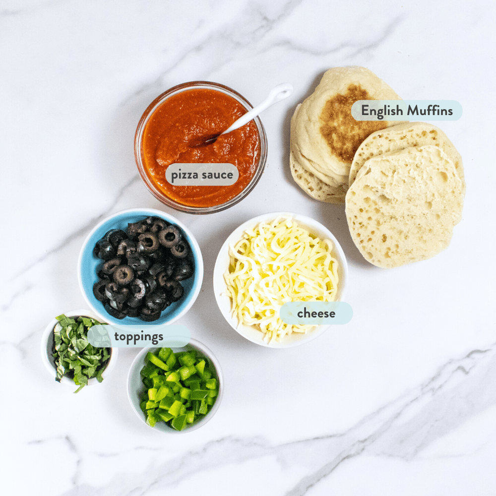 Spread on marble countertop of ingredients for English muffin pizzas including sauce, cheese, toppings and English muffins.