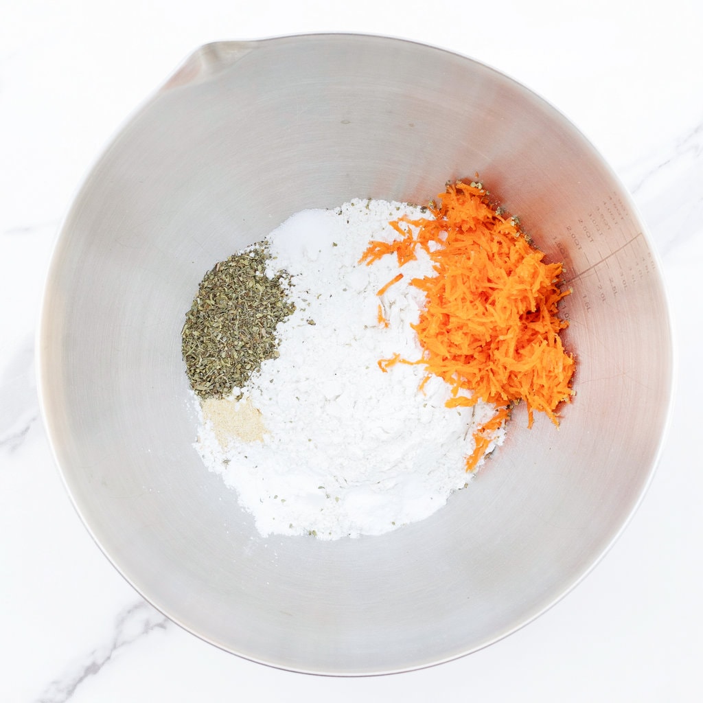 Silver mixing bowl on a marble countertop with dry ingredients for pizza muffins including carrots.