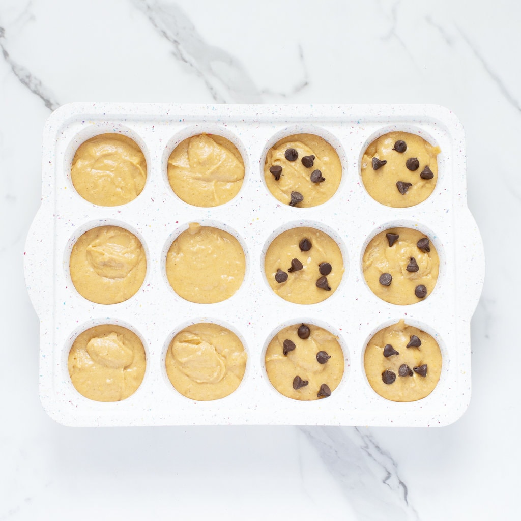 White muffin tin with wet muffin batter, some with toppings ready for the oven.