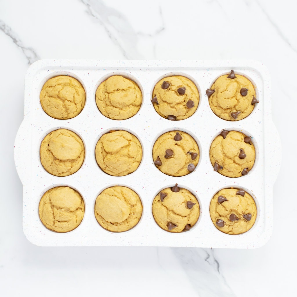 Sweet Potato muffins in a white muffin tray sitting on a marble countertop. Half of the muffins have chocolate chips are half of them do not.