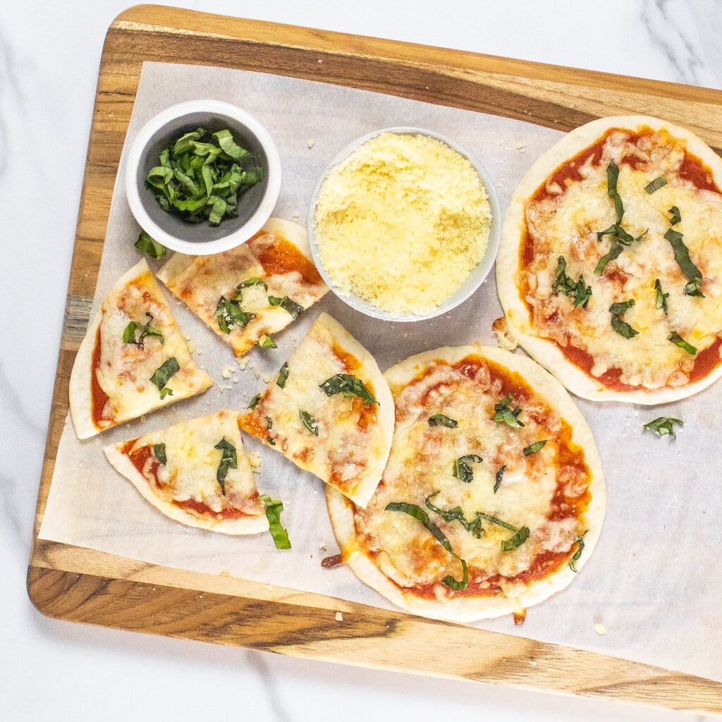 Wood cutting board on top of a marble countertop with tortilla pizza is cut on top with a small bowl of Parmesan and sliced basil.