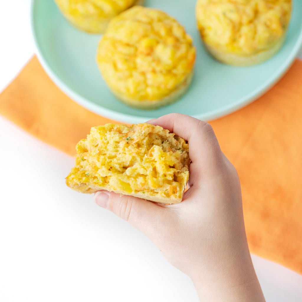 A kids hand holding a veggie muffin torn in half against a background of a blue kids plate and orange napkin.