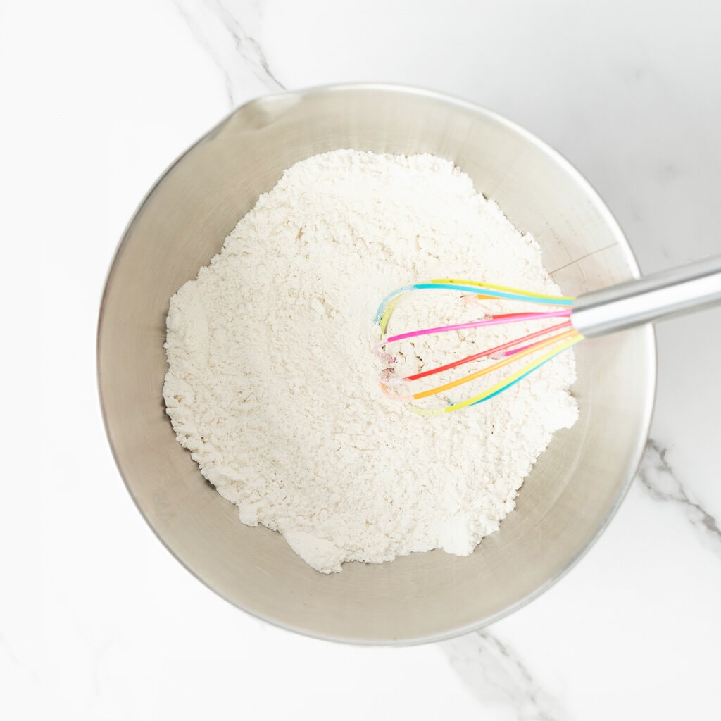 A silver mixing bowl with flour and dry ingredients for muffins on a marble countertop.