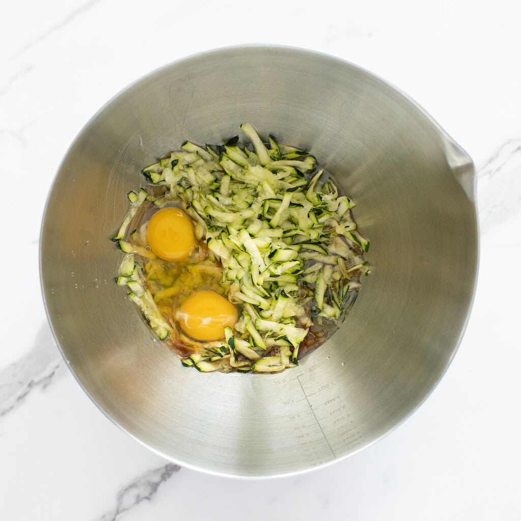 Silver mixing bowl on a marble counter filled with zucchini and other wet ingredients.