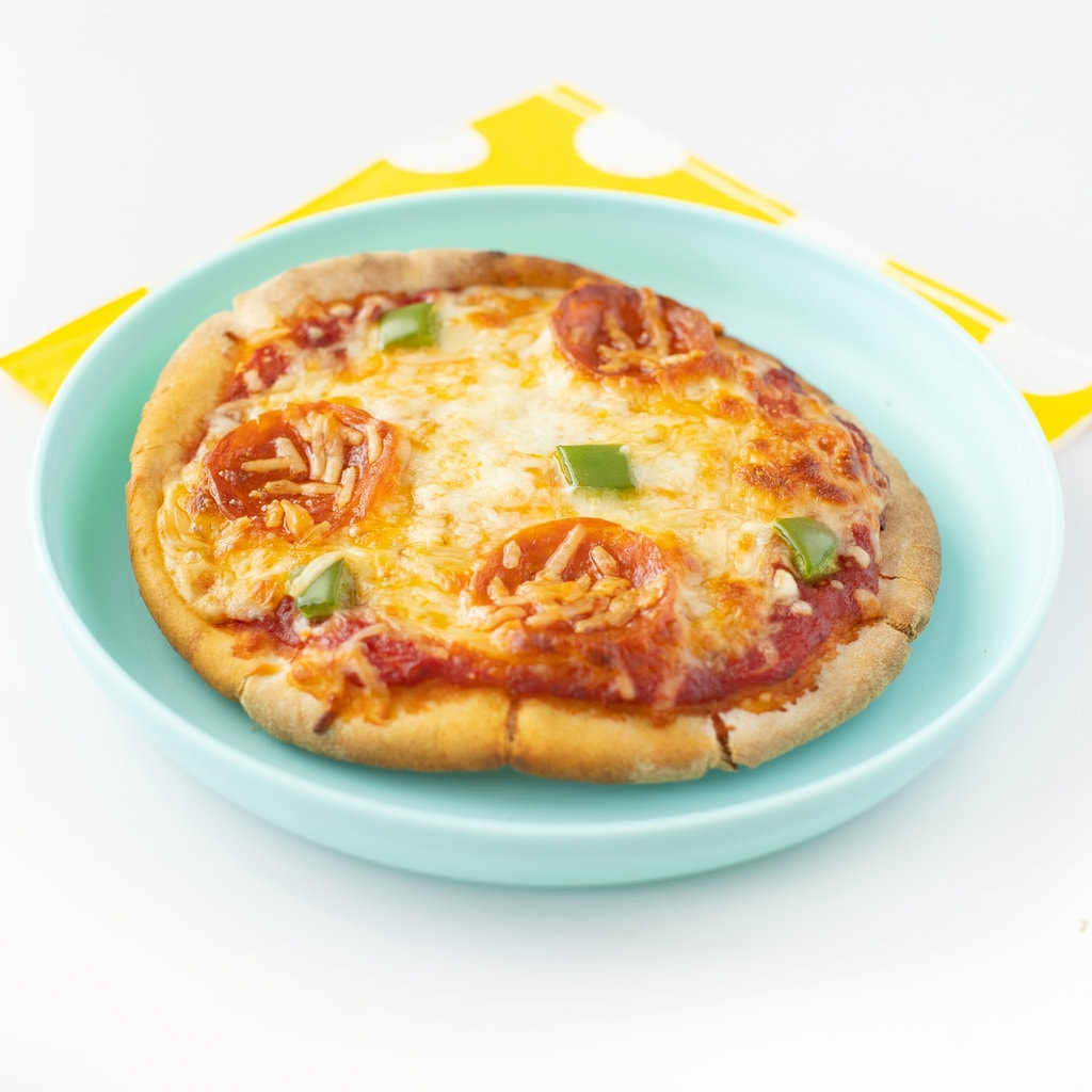 A pita pizza on a teal plate with a yellow napkin against a white background.
