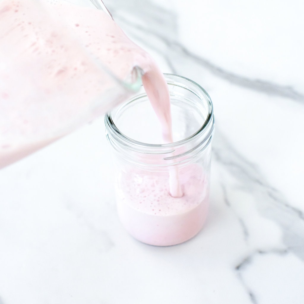 Clear blender pouring strawberry milk inside a clear class on a marble white counter.