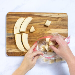 Two hands placing banana slices into a Ziploc bag off of a wood cutting board.
