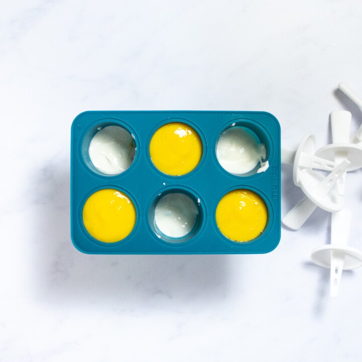 A dark teal popsicle molds with three of the molds filled with mango purée and the other three filled with yogurt with popsicle sticks sitting off to the side.