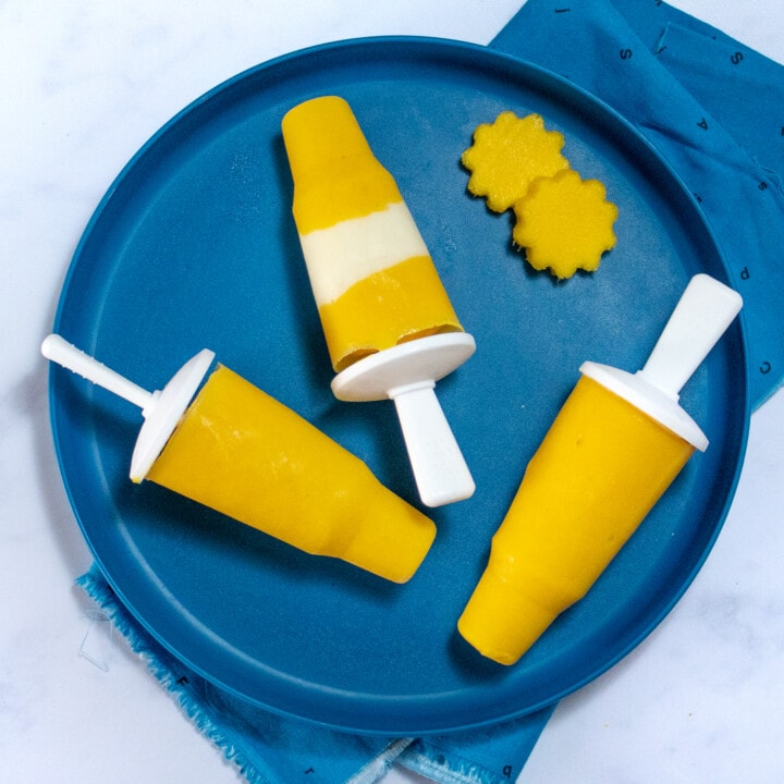 A dark blue plate full of three mango popsicles and a cut out with mangos.