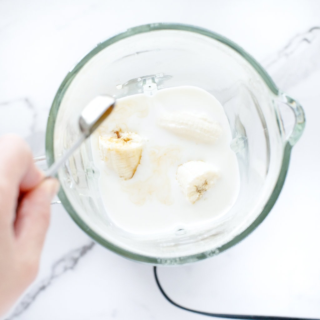 A clear blender with bananas and a hand pouring in Maple syrup.