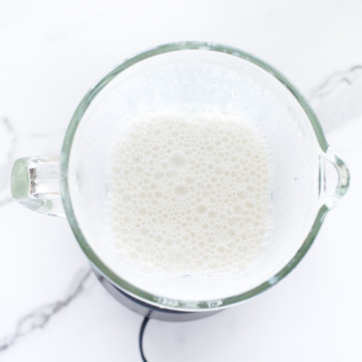 A clear blender full of milk against a white marble back ground countertop.