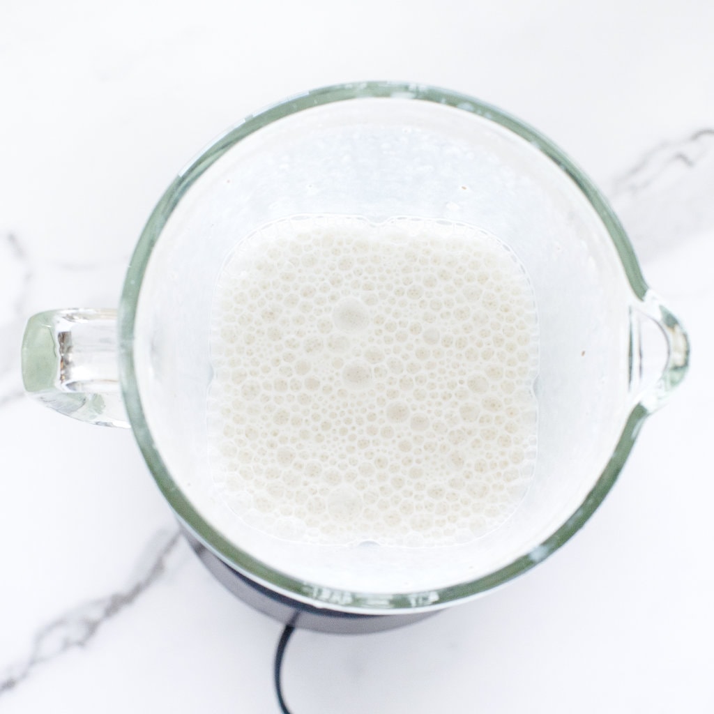 A clear blender full of milk against a white marble back ground countertop.