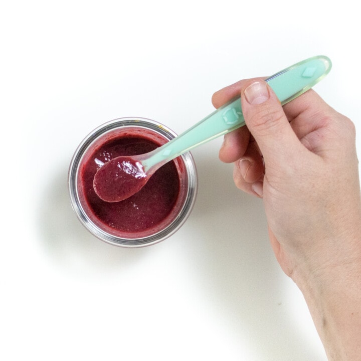 A clear glass baby food jar full of sod blackberry purée against a white background.