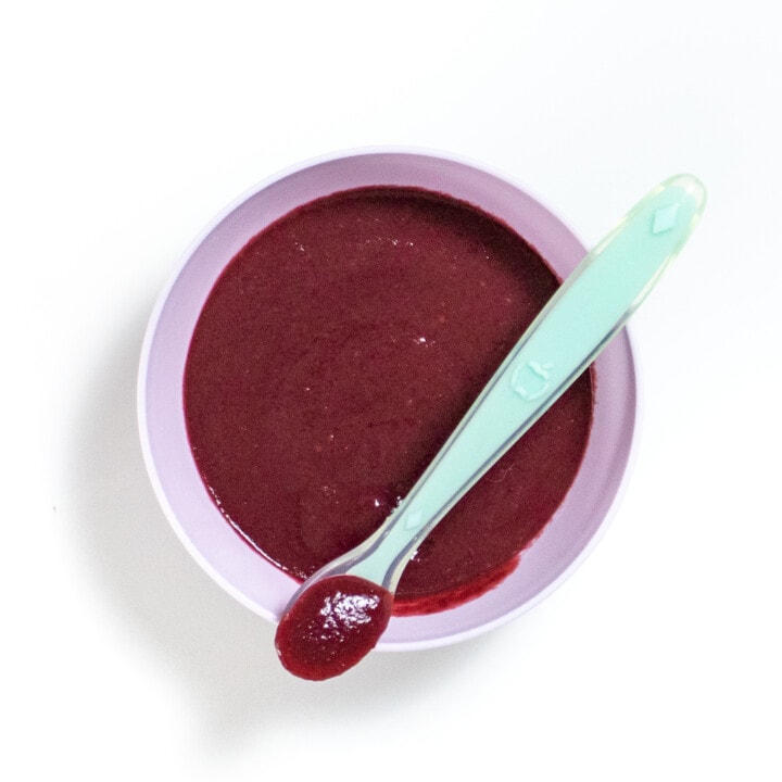 Purple baby bowl full of blackberry puree with a teal spoon resting on top.